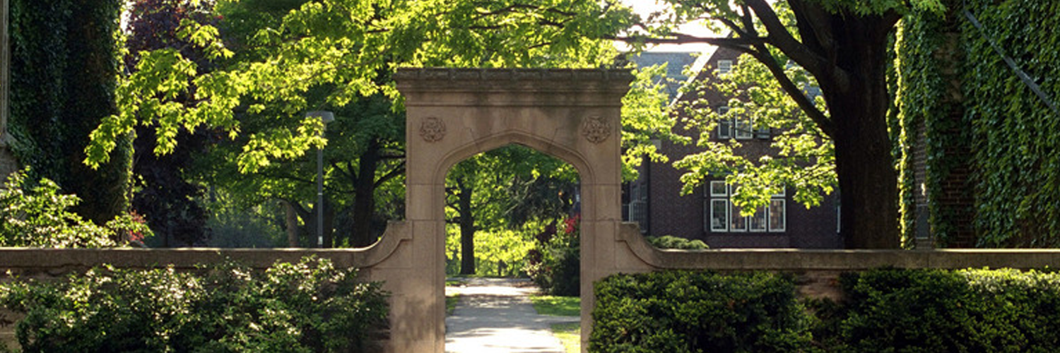 Stone archway in front of leafy trees and a brick building on the McMaster campus